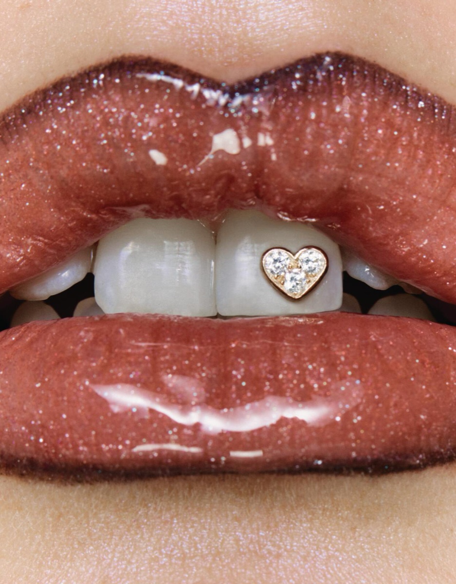 Close-up of glossy red lips parted to reveal a small heart-shaped diamond tooth jewel.