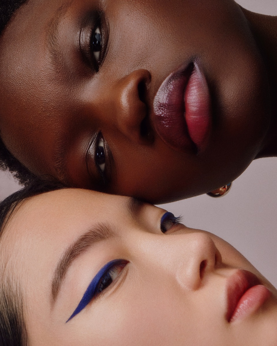 Two models' faces pressed together, one wearing bold blue winged eyeliner and the other with deep berry lipstick.